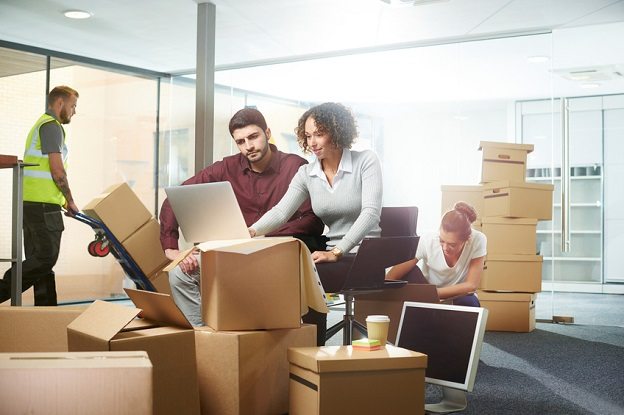 Three employees sitting in open office space surrounded by moving boxes and mover pushing boxes in a hand truck